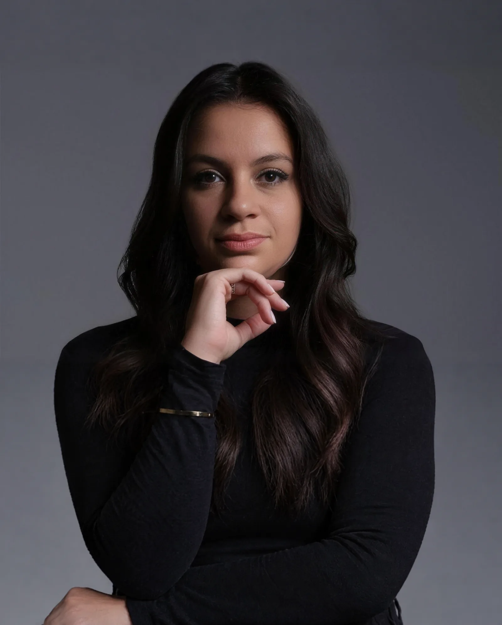 Dra. Débora Moraes em retrato de estúdio, de blusa preta, com a mão apoiada no queixo